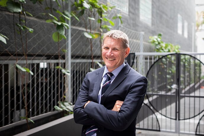 A man smiling with his arms folded and wearing a suit standing in front of a gate.