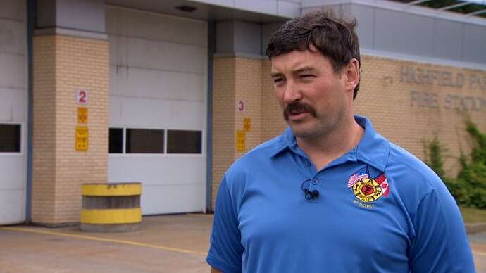 A man in a blue firefighter union shirt stands in front of a fire station.