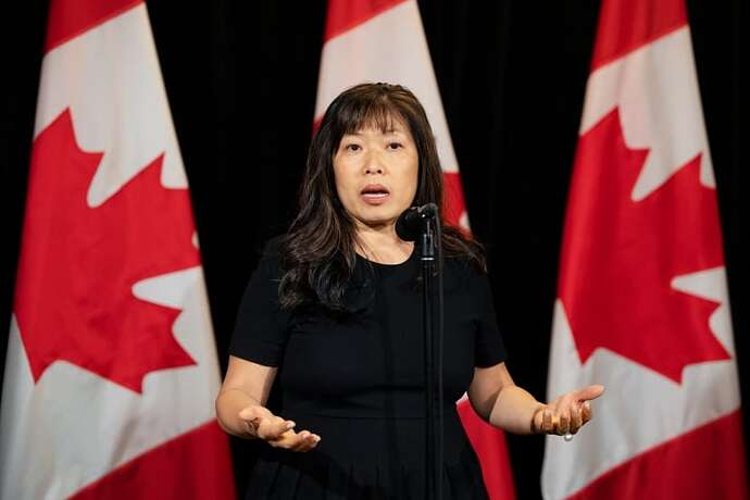 A woman in a dark dress speaks at a microphone with Canadian flags hanging in the background.
