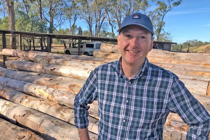 A man wearing a blue cap and a blue-and-white checked shirt stands in front of timber logs.