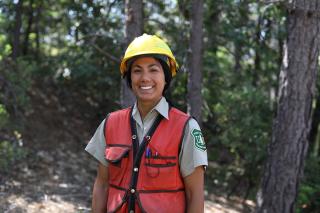 Sonya Lucatero wearing a hard hat, high visibility jacket, over a USDA Forest Service uniform, standing in a forest.
