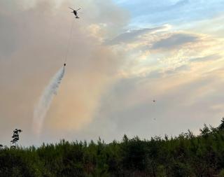 A helicopter drops water from a suspended bladder onto the burning forest below.
